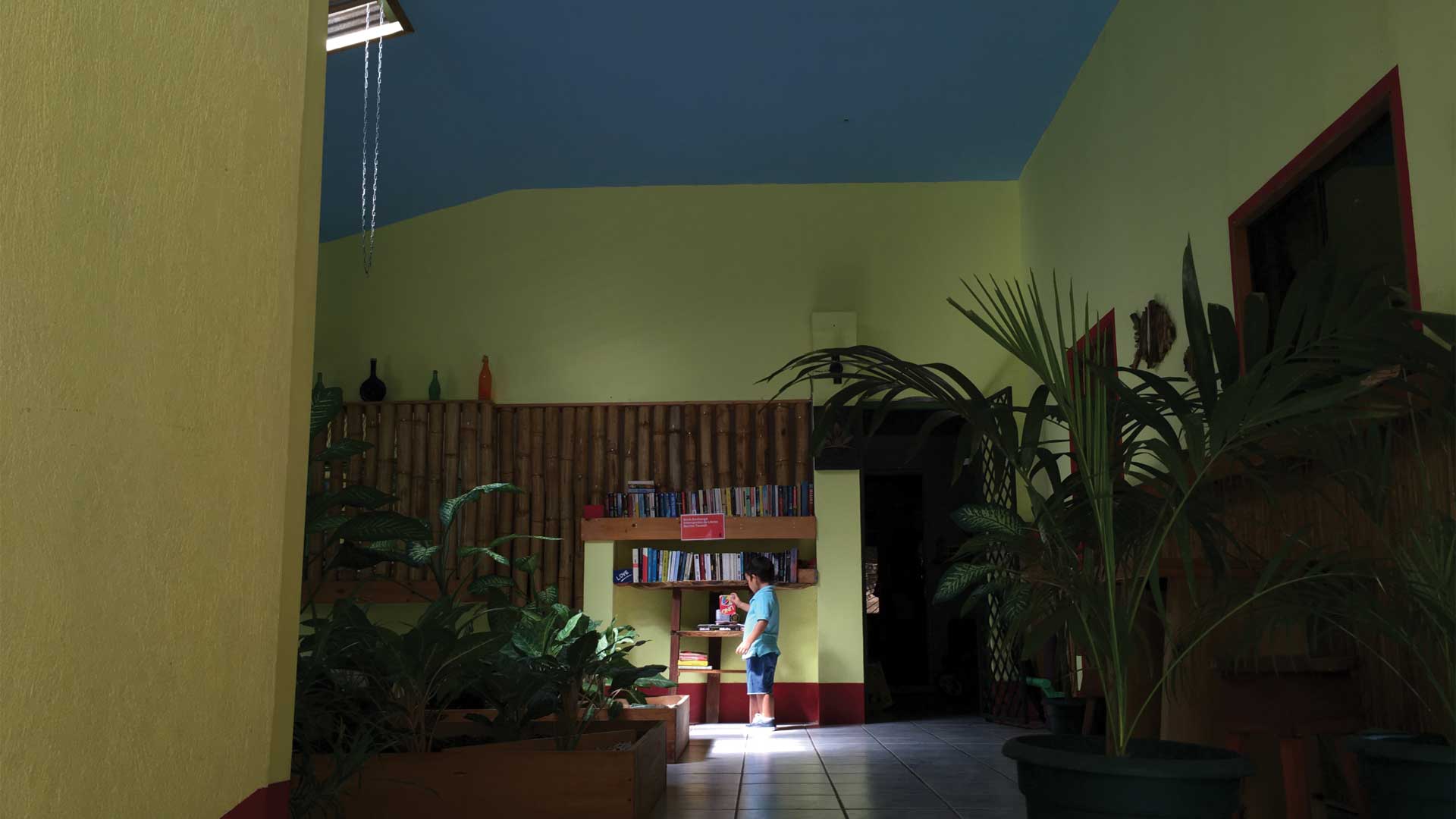 Boy picks up item at bookshelf in corridor by indoor gardens of boutique hotel Indra Inn at Playa Grande, Costa Rica