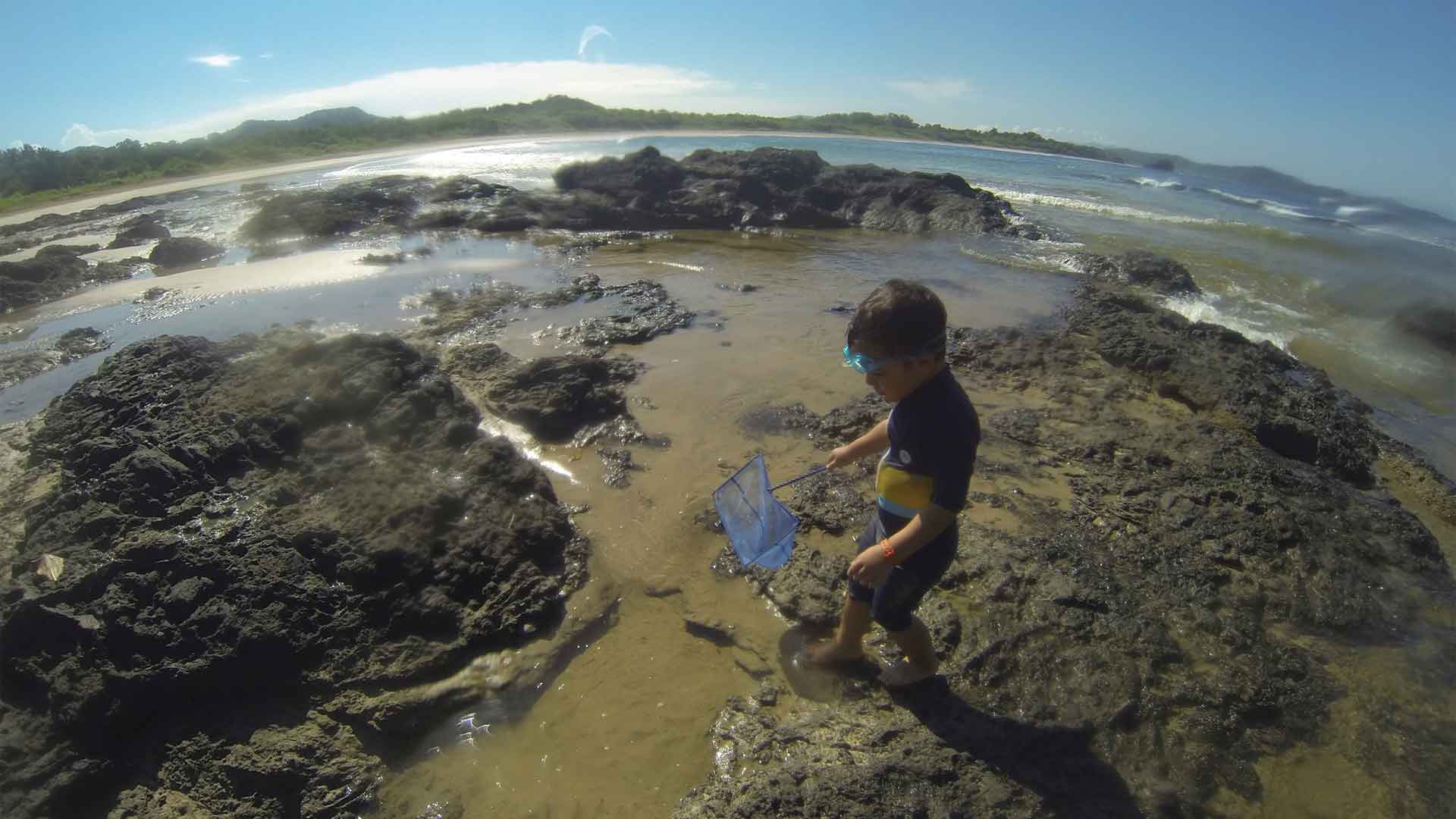 Boy in wetsuit walking with net and goggles over mossy black rock through tide pools at Playa Carbon beach in Guanacaste, Costa Rica