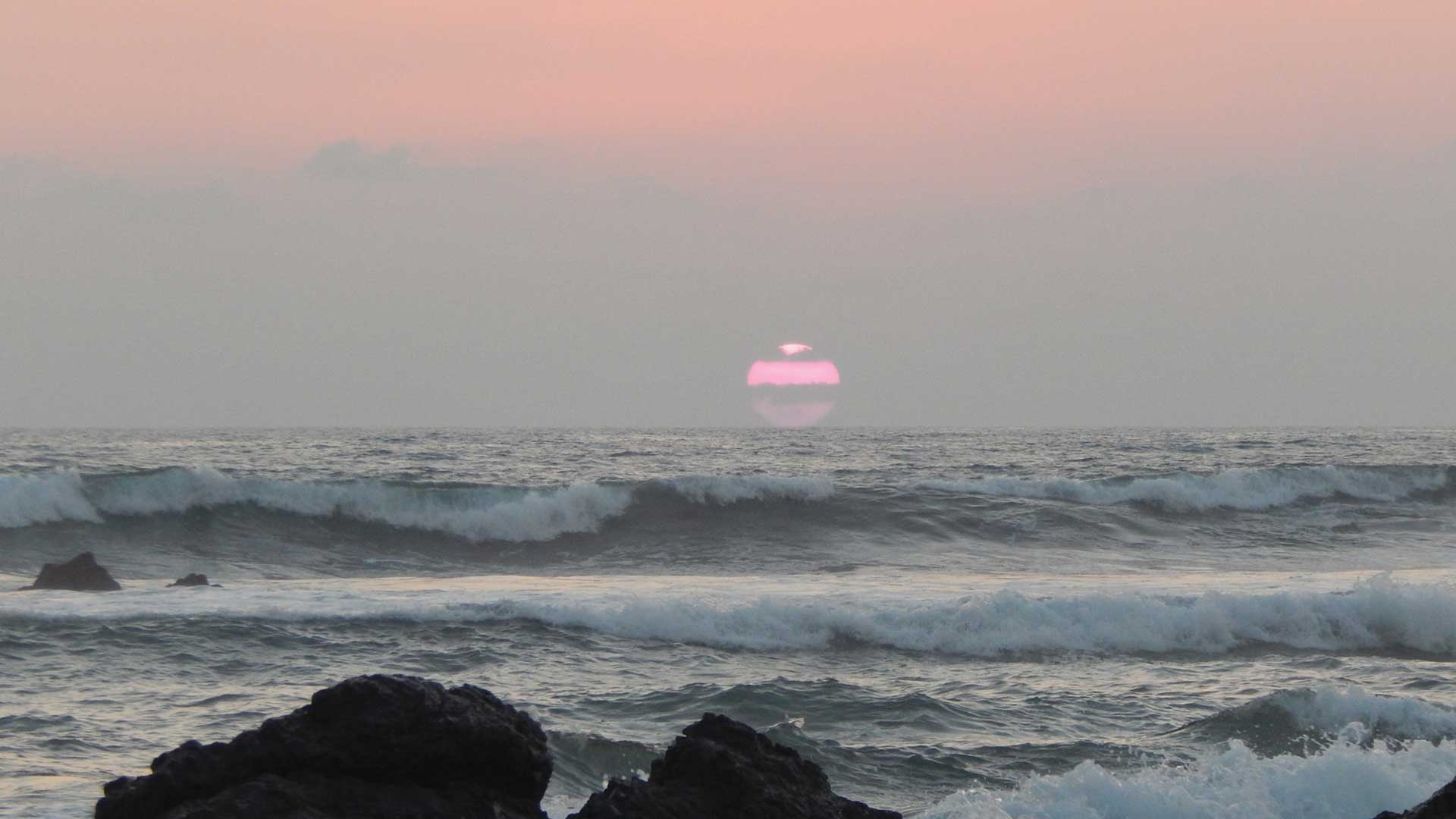 Large red sun hazy sunset with waves crashing onto black rock at Playa Grande beach in Guanacaste, Costa Rica